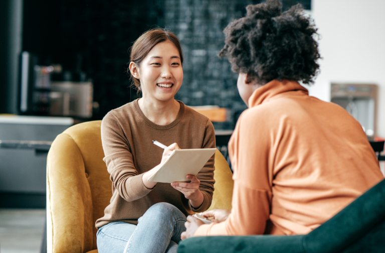 A smiling counselor holding a clipboard talks with a client seated across from her in a comfortable, casual setting.