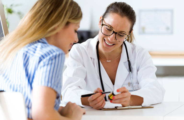 A female doctor in a white coat, wearing glasses and a stethoscope, smiles while discussing medication with a patient in a striped shirt across the desk.