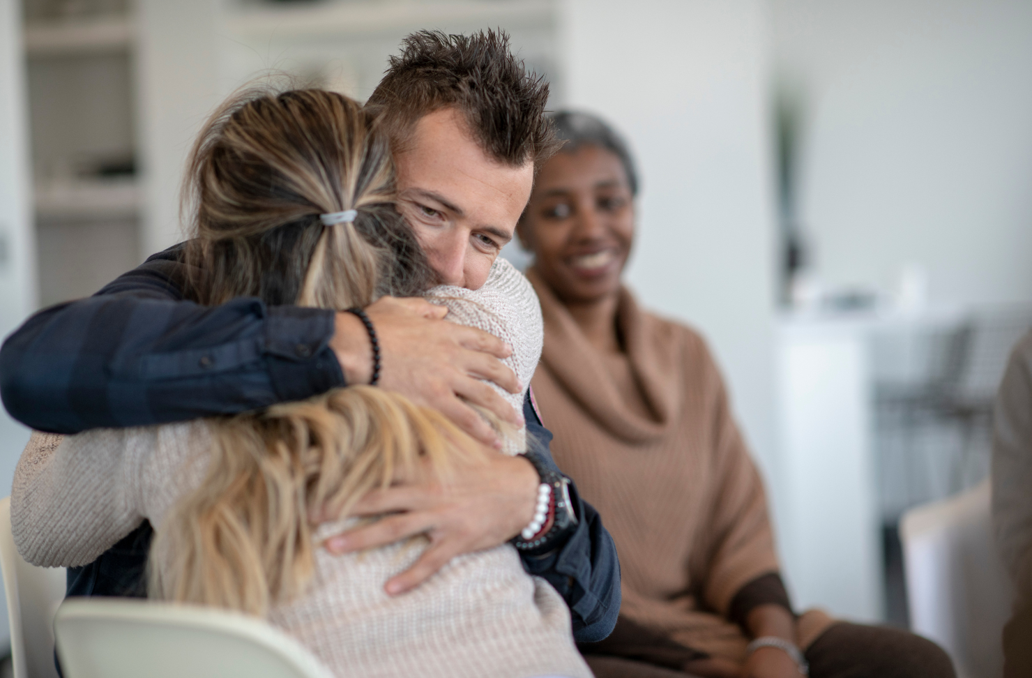 A man warmly hugs a woman during a support group session while another participant smiles in the background.