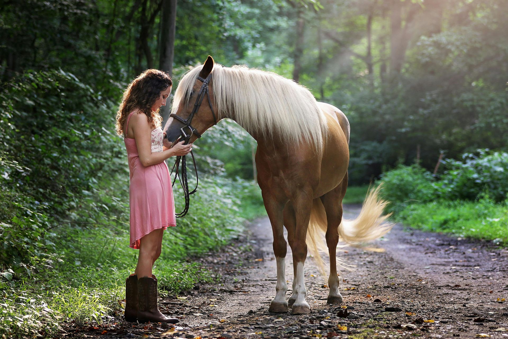 A woman in a pink dress is standing next to a brown horse on a dirt road.