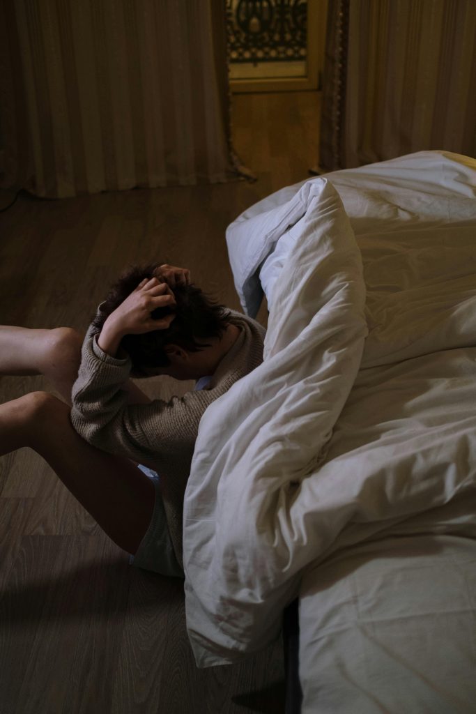 Man sitting on the floor next to a bed with a white comforter struggling with methamphetamine addiction.