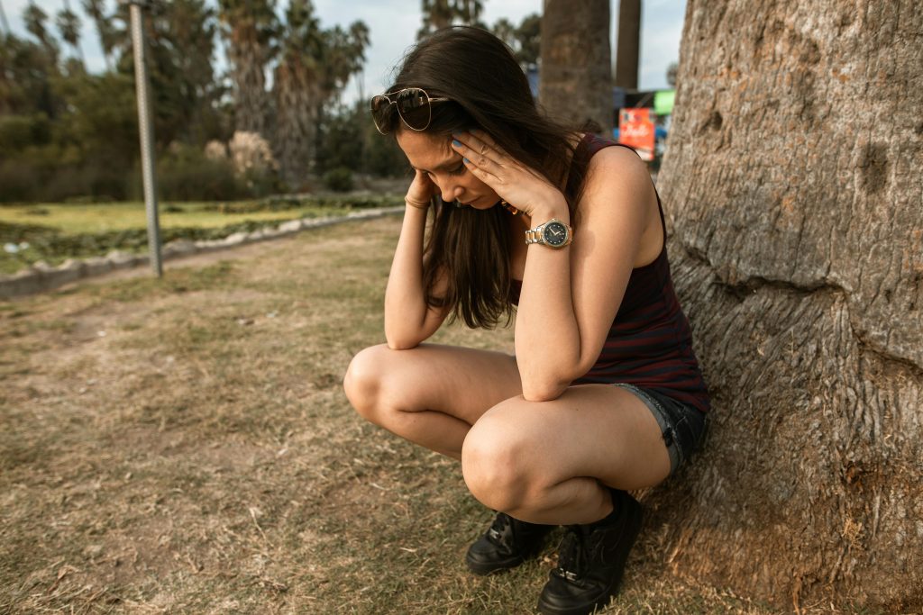 Woman wearing a tank top and shorts sitting next to a tree with her hands on her head struggling with methamphetamine addiction in Los Angeles.