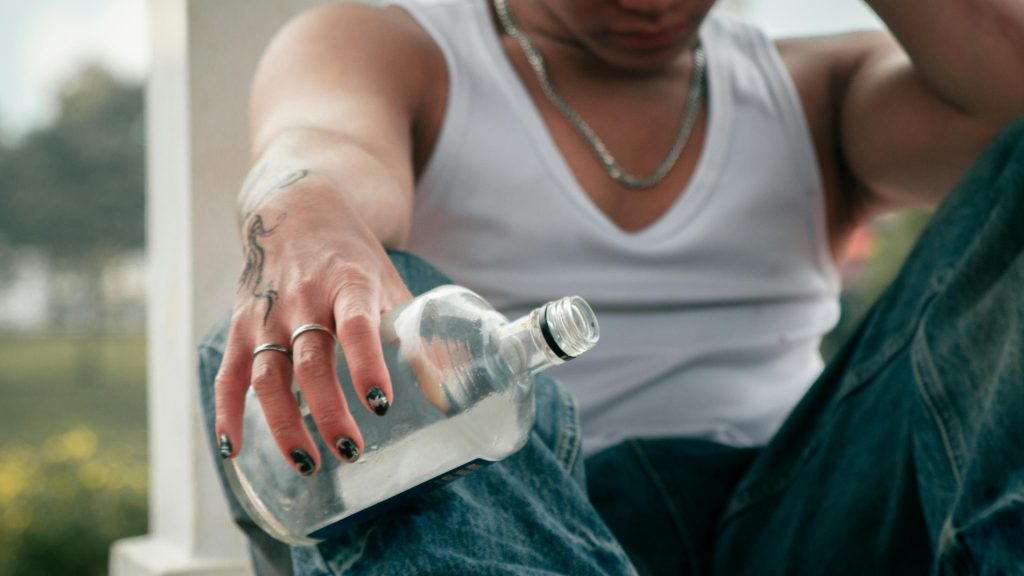 Close up of a man holding an empty glass bottle with one hand on his forehead showcasing the downfall and turmoil of alcohol addiction.